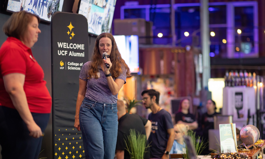 Woman with a microphone speaking on stage at a UCF Alumni welcome event; banner reads 'WELCOME UCF Alumni' behind her.