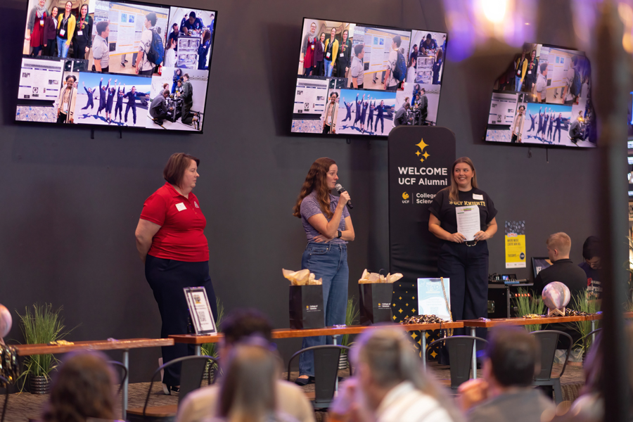 Three women on a stage at a UCF Alumni event; one speaks into a microphone while another holds a certificate, with screens showing photos in the background.