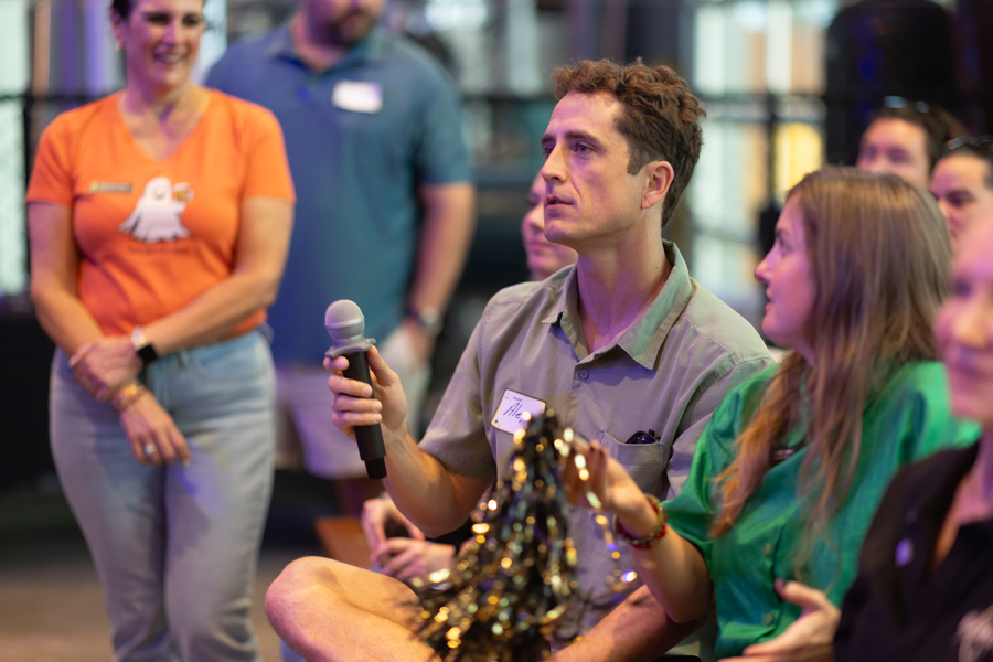 Man in a gray shirt holds a microphone while seated, surrounded by attentive attendees at a casual event.