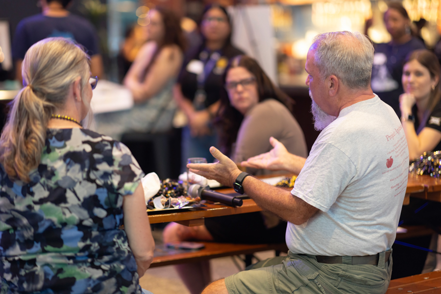 Older man with a gray beard in a light T-shirt talks to a woman at a crowded event table surrounded by onlookers.