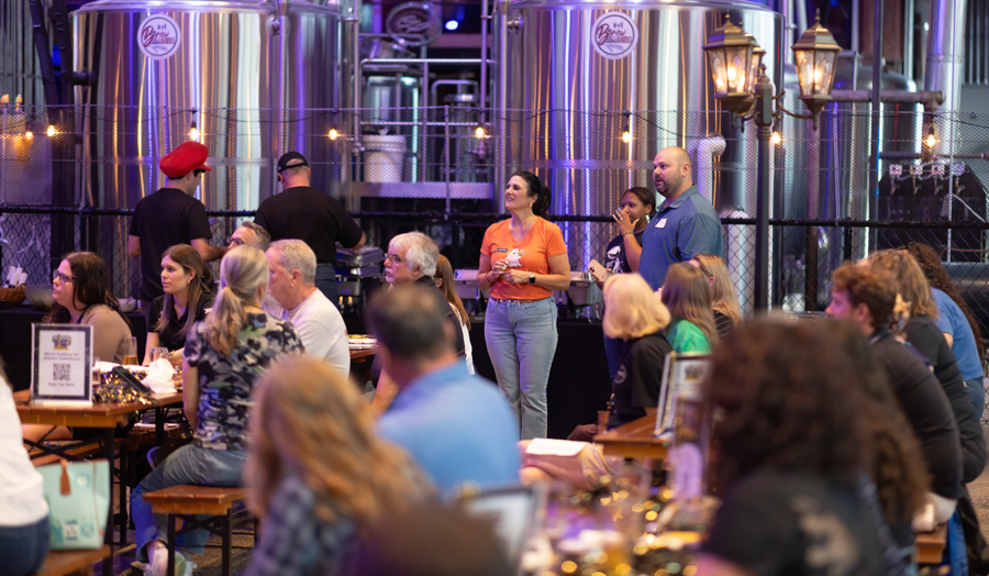 Group of people seated at wooden tables in a brewery tasting event, with large stainless tanks in the background.
