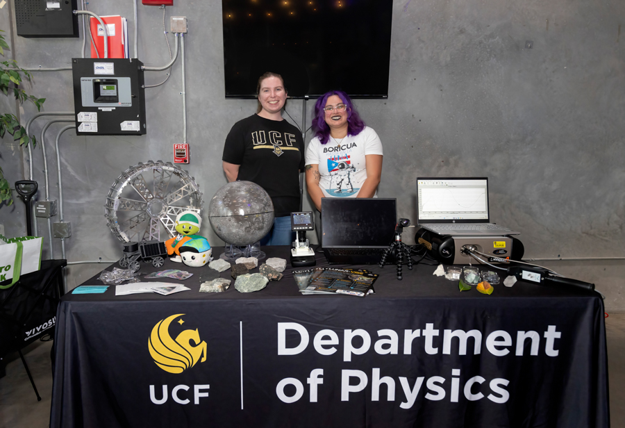 Two people stand behind a UCF Department of Physics table with science demos and equipment on display, smiling at the camera.