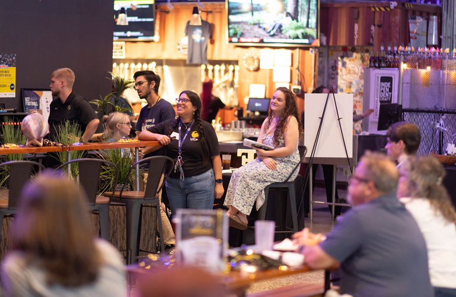 Group of people gathered at a bar-like counter, some standing and others seated, watching something off-camera; a woman sits on a stool with a notebook, smiling.