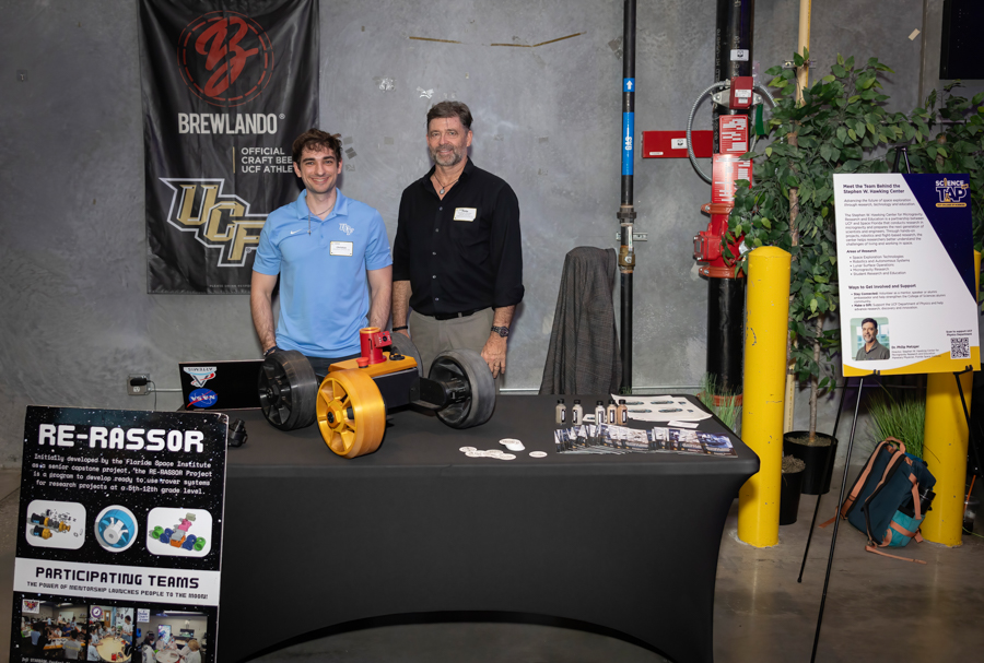 Two men stand behind a black display table at a science expo, presenting a yellow wheeled rover with its components laid out and brochures nearby.