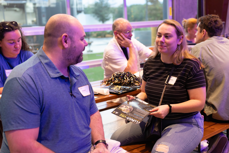 Two adults seated at a wooden table in a bright, modern space chat; the woman holds a flyer about the event while other people talk in the background.