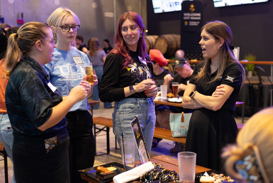 Four women standing around a table, chatting and holding drinks at a social event in a lively venue.