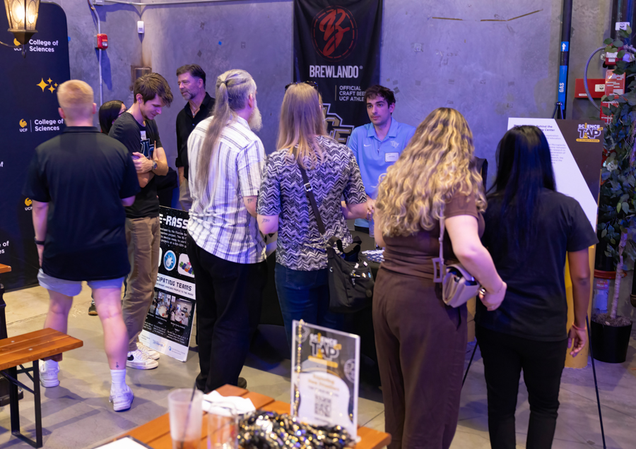 Group of people gathered around a booth at a science‑expo/event; banners for Brewlando and College of Sciences visible in the background.