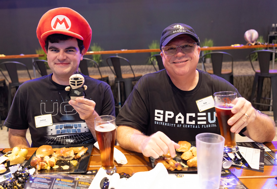 Two men sit at a table with sliders and beer, one wearing a red Mario hat and holding a small figurine, both smiling at the camera.