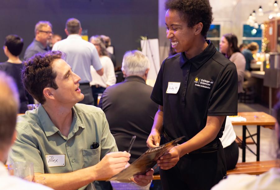 Man named Alex signs a form at a table as a student ambassador in a black polo hands him a brochure at a college event reception.