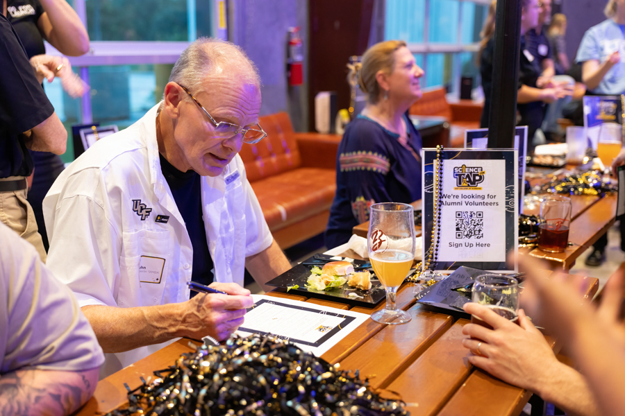 Older man in a white lab coat signs a form at a bar-style table during a science outreach event, with snacks and drinks nearby and a sign-up sheet visible for alumni volunteers (Science Tap).