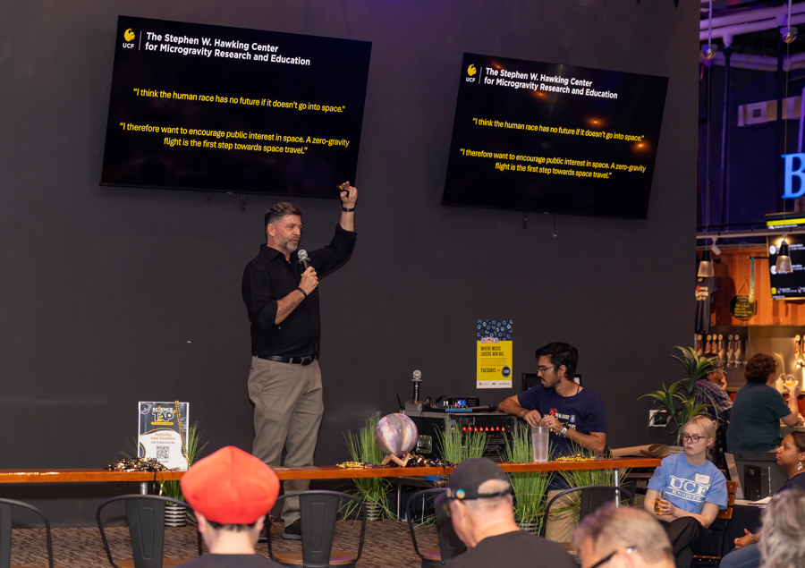 Male speaker on a dim stage, microphone in hand, raising a fist as he talks; two screens above show yellow quotes about space.