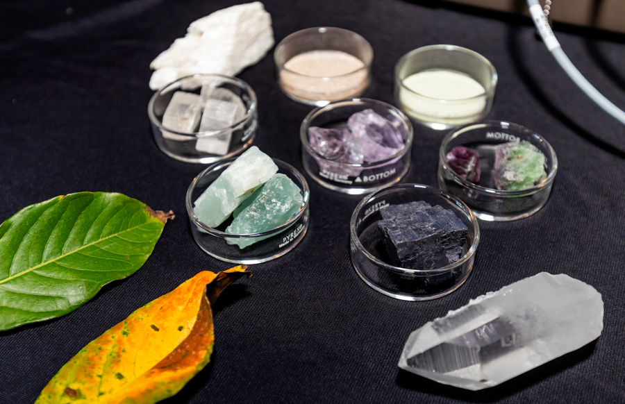 Assorted mineral and crystal samples in glass containers on a dark cloth surface, with green leaves nearby.