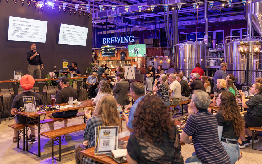 Speaker on stage presenting to a seated crowd at a brewery event, with large screens and brewing tanks in the background