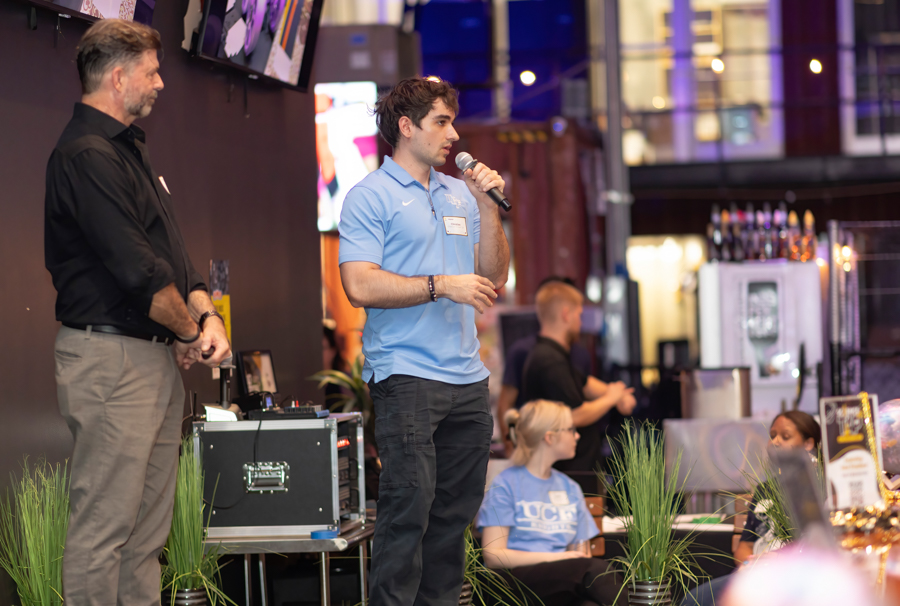 Man in a light blue polo speaks into a microphone on a stage at an indoor event, with a second man standing nearby and equipment behind them.
