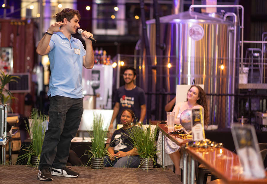 Male host in a light blue polo speaks into a microphone on stage at a brewery event, audience watching nearby.