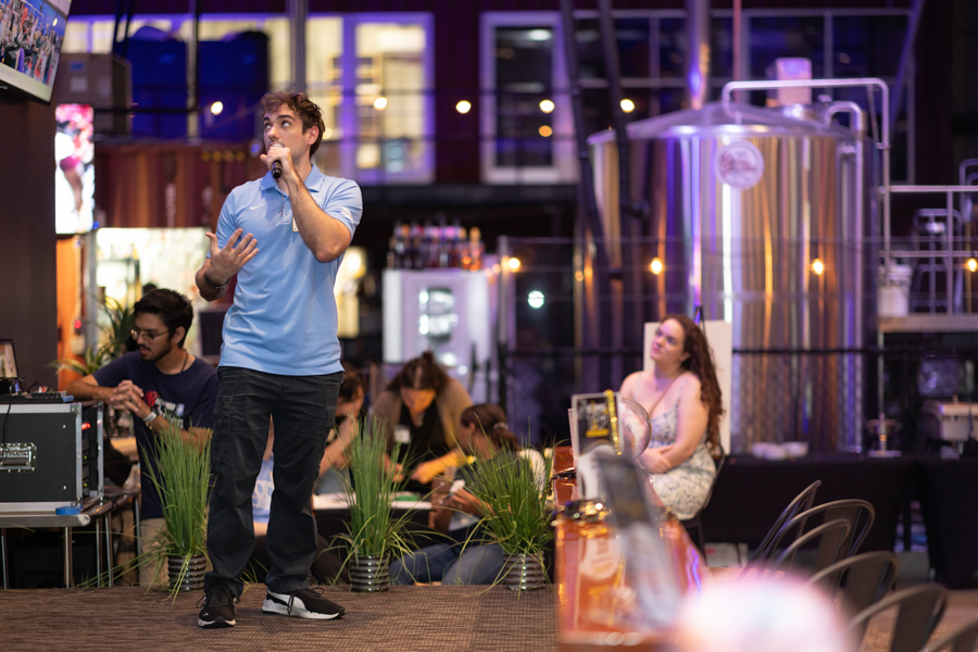 Man in a light blue polo sings into a microphone on a stage at an indoor event, with equipment and attendees in the background.