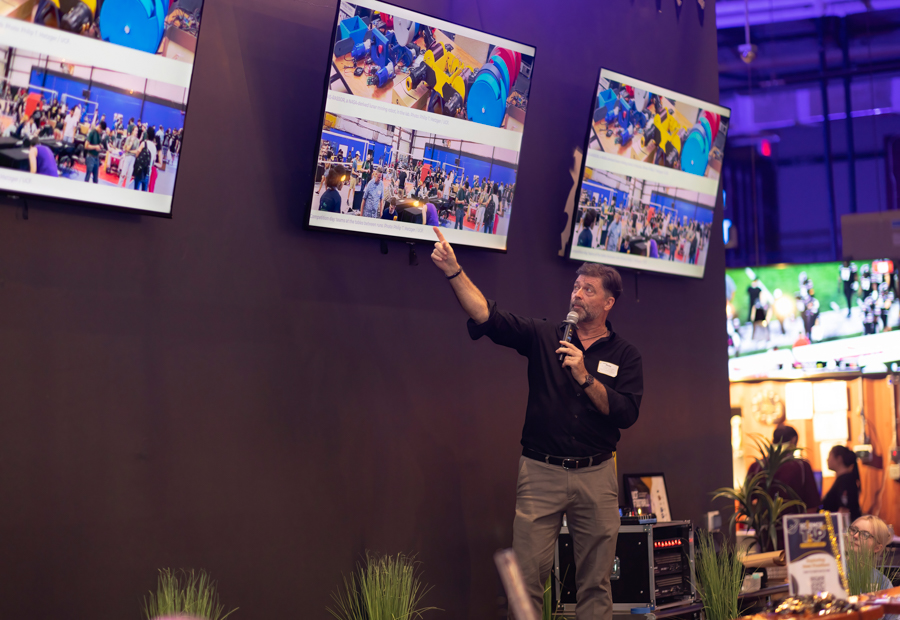 Male presenter with microphone pointing at three mounted screens showing a crowded trade show room.