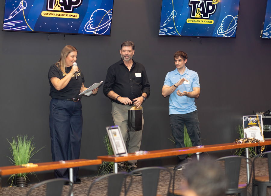Woman speaks into a microphone while holding papers on a stage, flanked by two men; one holds a gift bag.