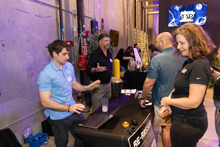 Young man in a light blue polo demonstrates a laptop at a tech booth to a small group at an indoor event venue.