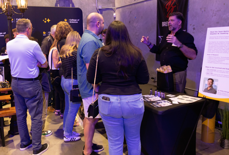 A presenter speaks to a line of attendees at a science expo booth, with College of Sciences banners in the background.