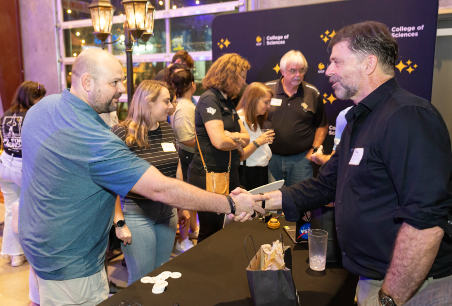 Two men shake hands at a booth during a science college event, with attendees in the background and a 'College of Sciences' banner behind them.