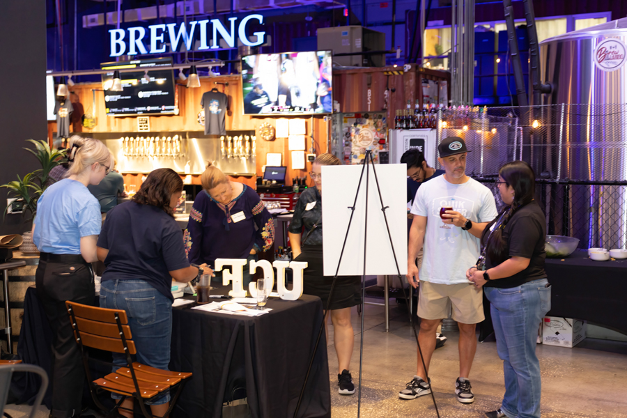 People gather around a brewery booth at an indoor beer festival, with a bright 'BREWING' sign overhead and a white presentation board nearby.