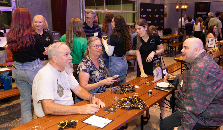 Group of people seated at a wooden table chatting and enjoying drinks at a social event; a server in black assists nearby.