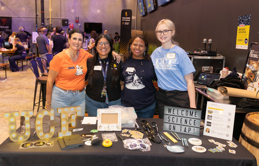 Four women pose together behind a UCF booth table at a science event, with UCF letters and science-related items on display.