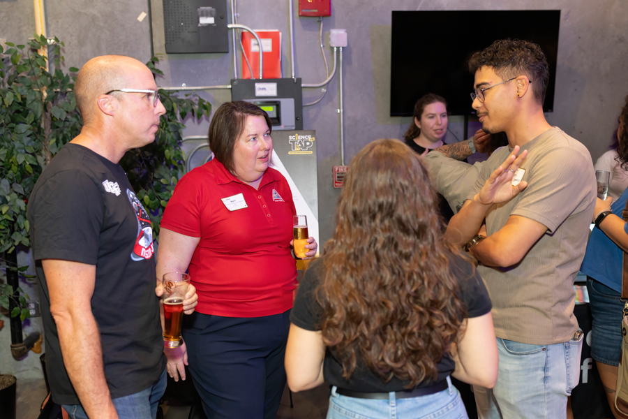 Group of adults chatting at an informal networking event, holding beer glasses indoors.