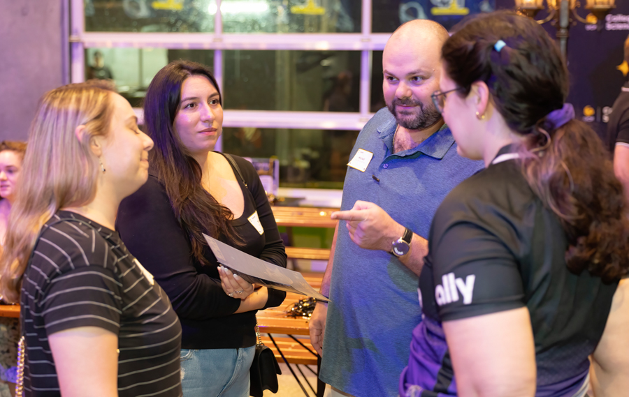 Four adults in a casual networking scene, a man points as two women hold papers and listen.