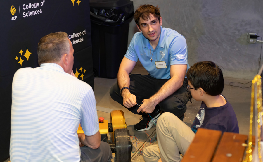 Instructor and two students crouch on the floor, wiring a small mechanical device at a College of Sciences event.