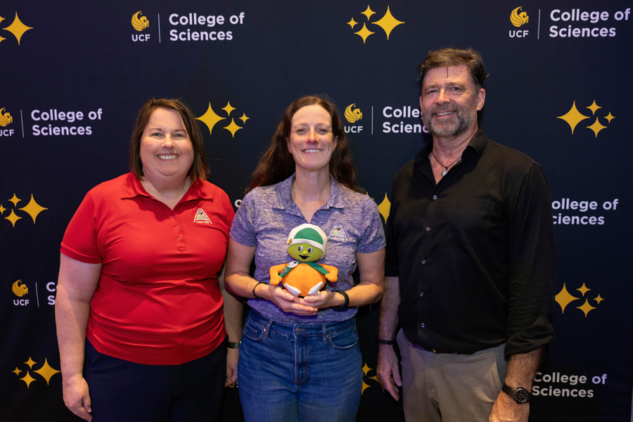 Three adults posing for a College of Sciences backdrop; middle person holds a plush mascot toy.