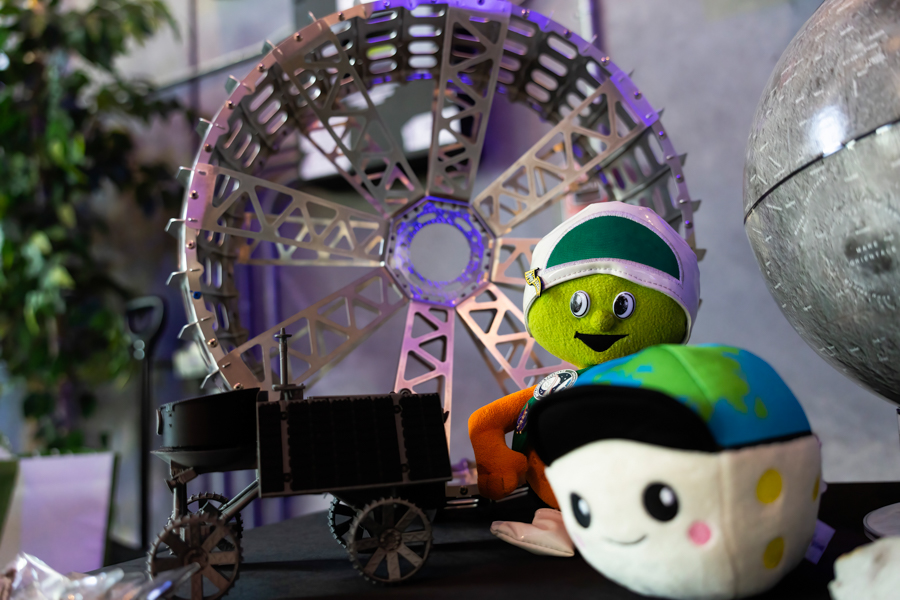 Metal lattice wheel structure in the background with a green plush astronaut and a globe plush on a display table.