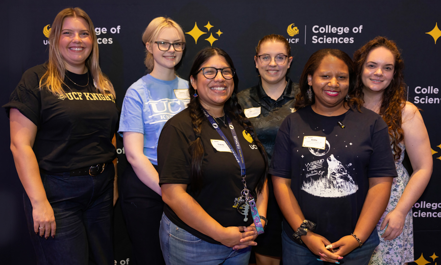 Six women stand together and smile for a group photo in front of a UCF College of Sciences backdrop; several wear name tags and UCF shirts.