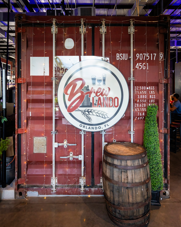 Circular Brew Orlando sign on a red shipping container, with beer icons and barley, a wooden barrel in front, in a brewery setting.