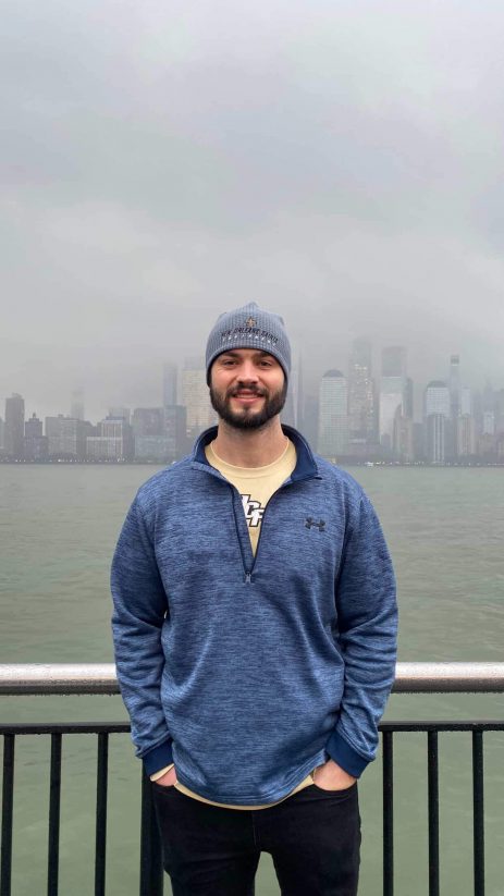 Smiling man in a blue pullover and beanie stands by a railing along the water with a foggy city skyline in the background.