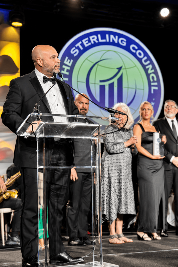 Man in a black tuxedo and bow tie speaks at a clear podium on stage, with a large 'The Sterling Council' emblem in the background and people watching behind him.