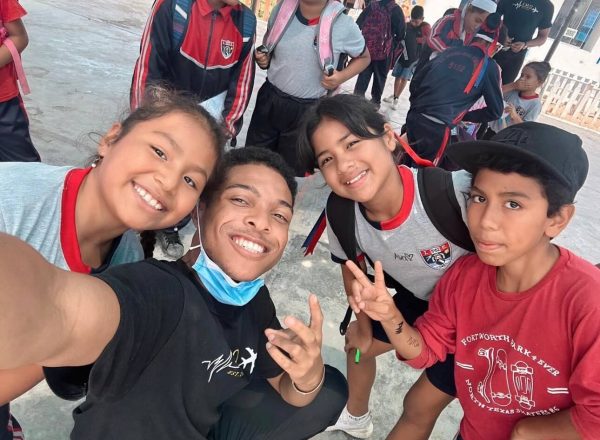 Group of smiling students and a man taking a selfie, outdoors on a school day in red and gray uniforms.