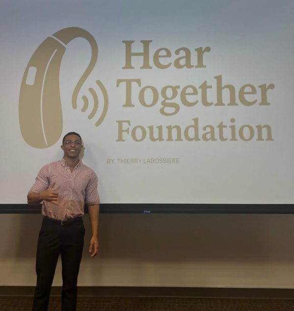 Man in a red-checkered shirt posing in front of a projected 'Hear Together Foundation' slide on a screen behind him.