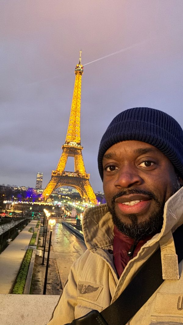 Selfie of a man in a beige jacket and navy beanie with the illuminated Eiffel Tower in Paris behind him at dusk/night.
