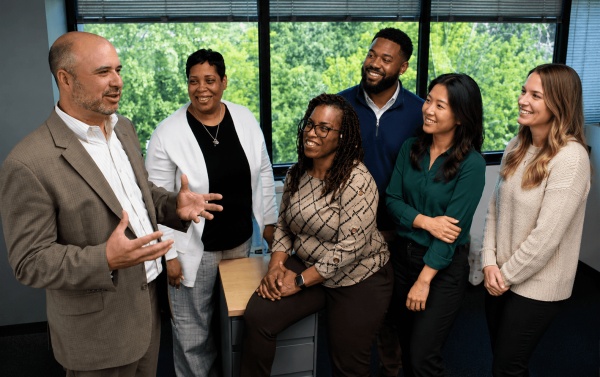 Six coworkers stand around a desk in a bright office as a man in a tan suit speaks, others smiling and listening.