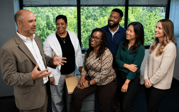 Six coworkers stand around a desk in a bright office as a man in a tan suit speaks, others smiling and listening.
